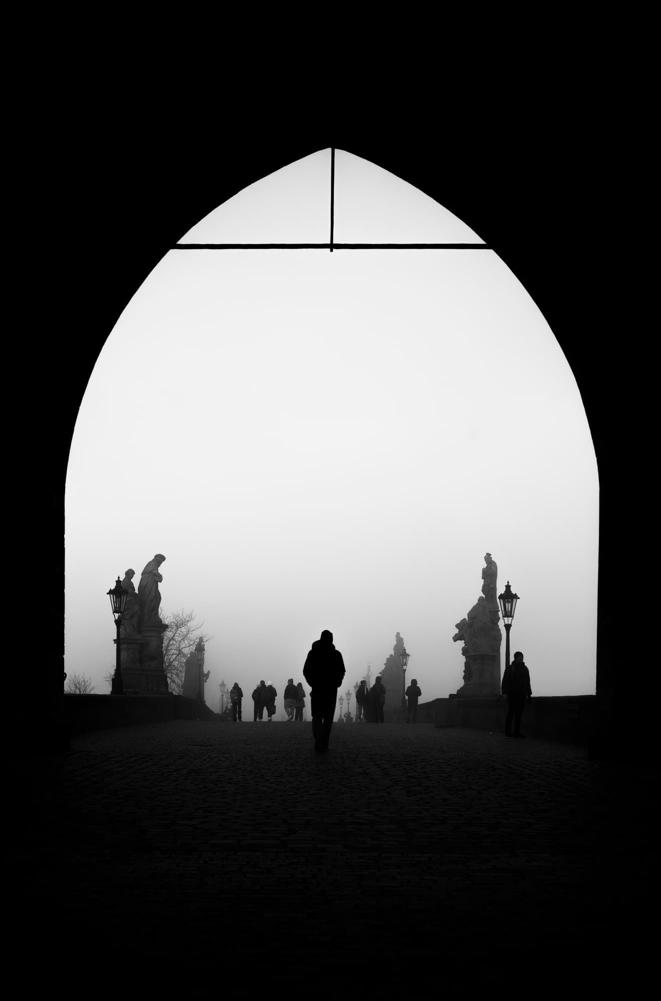 A silhouetted figure walking through Charles Bridge’s stone archway into thick fog, statues lining the way