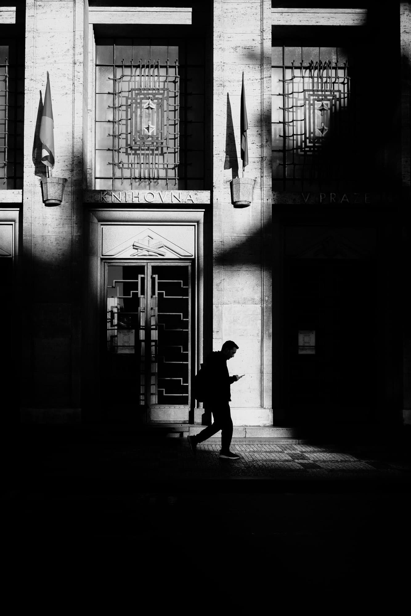 A lone figure crossing a blade of hard sunlight outside the Prague Municipal Library, deep shadow across the stone facade