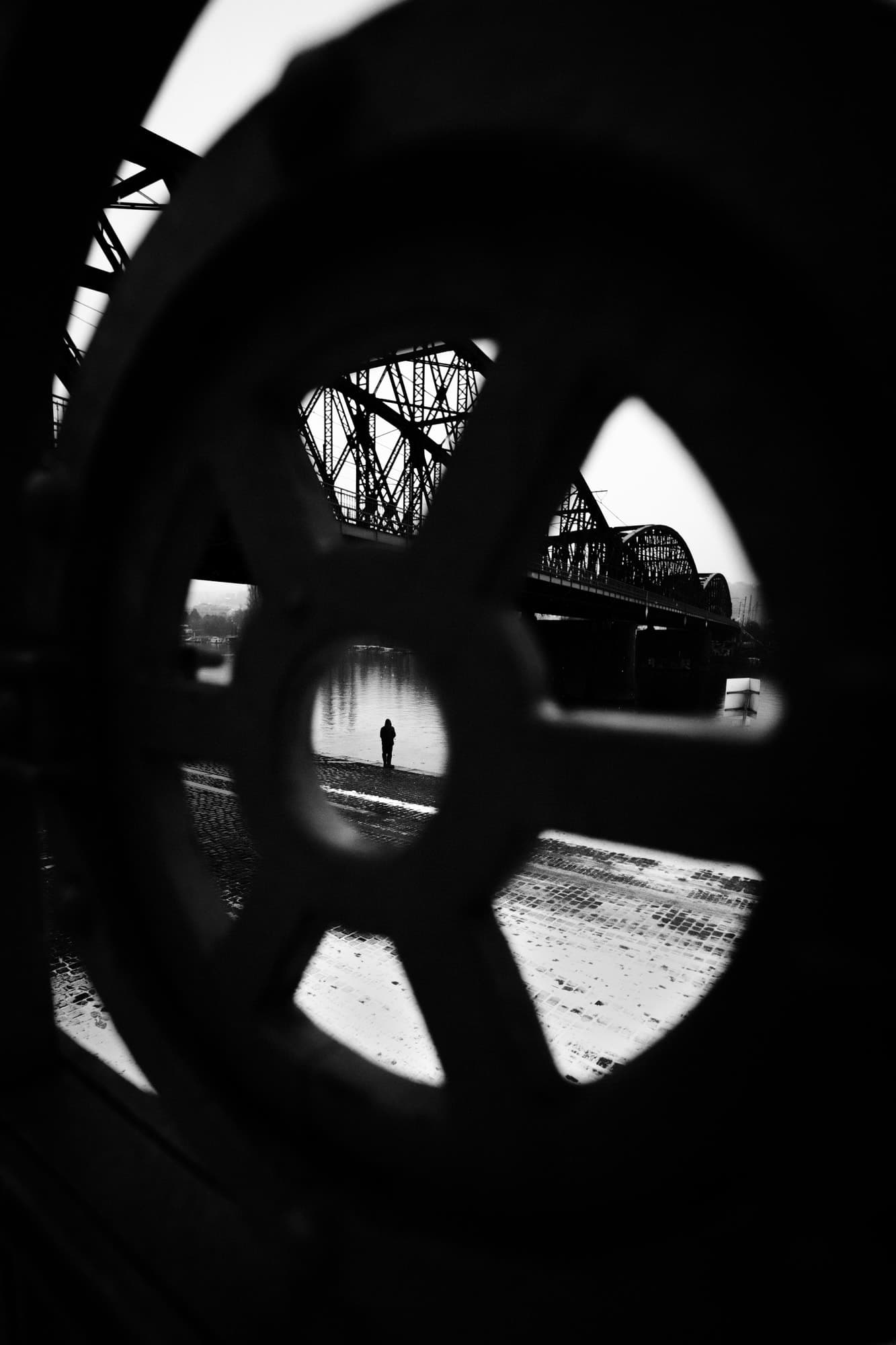 A solitary figure framed through the wheel of an old iron gate, a railway bridge beyond
