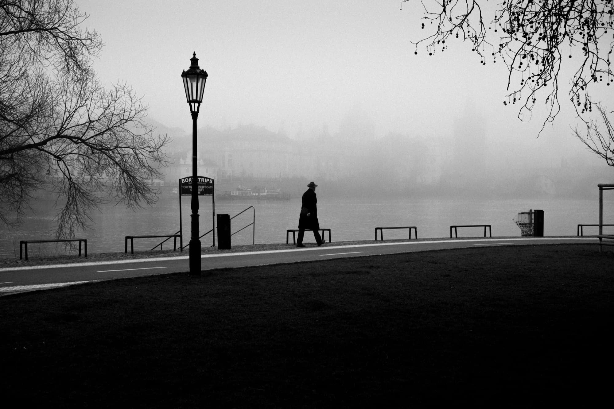 A lone figure in hat and coat on a foggy riverside promenade, a lamppost beside them